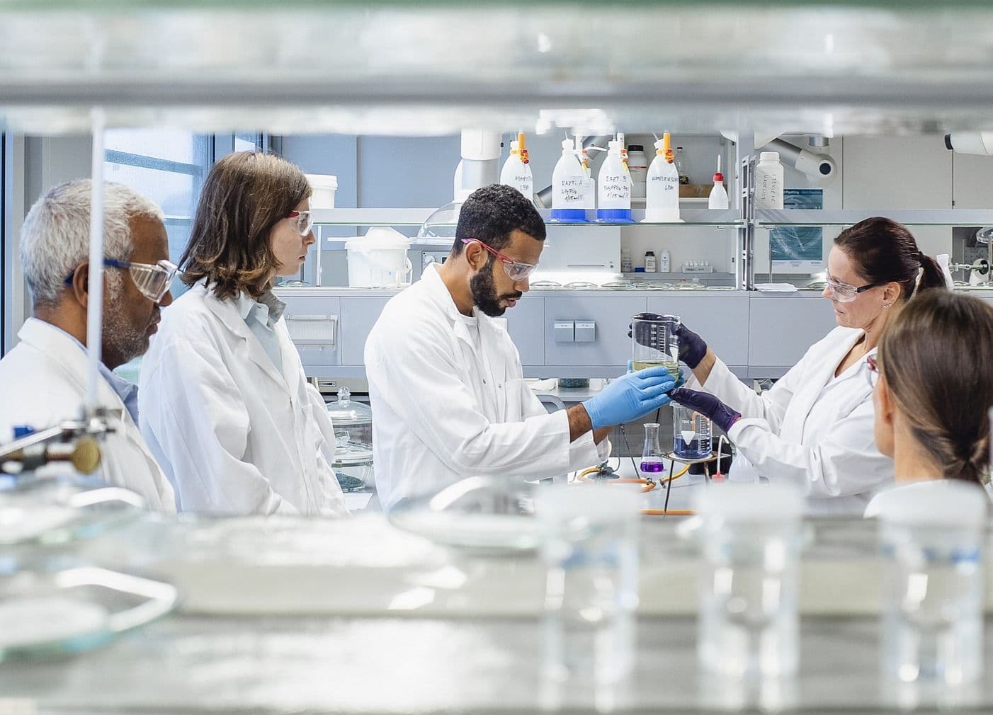 Team of scientists in lab coats and safety glasses collaborate around a bench, examining a beaker