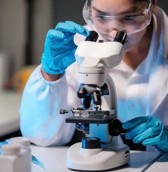 Scientist in safety goggles and blue gloves examining a sample through a laboratory microscope