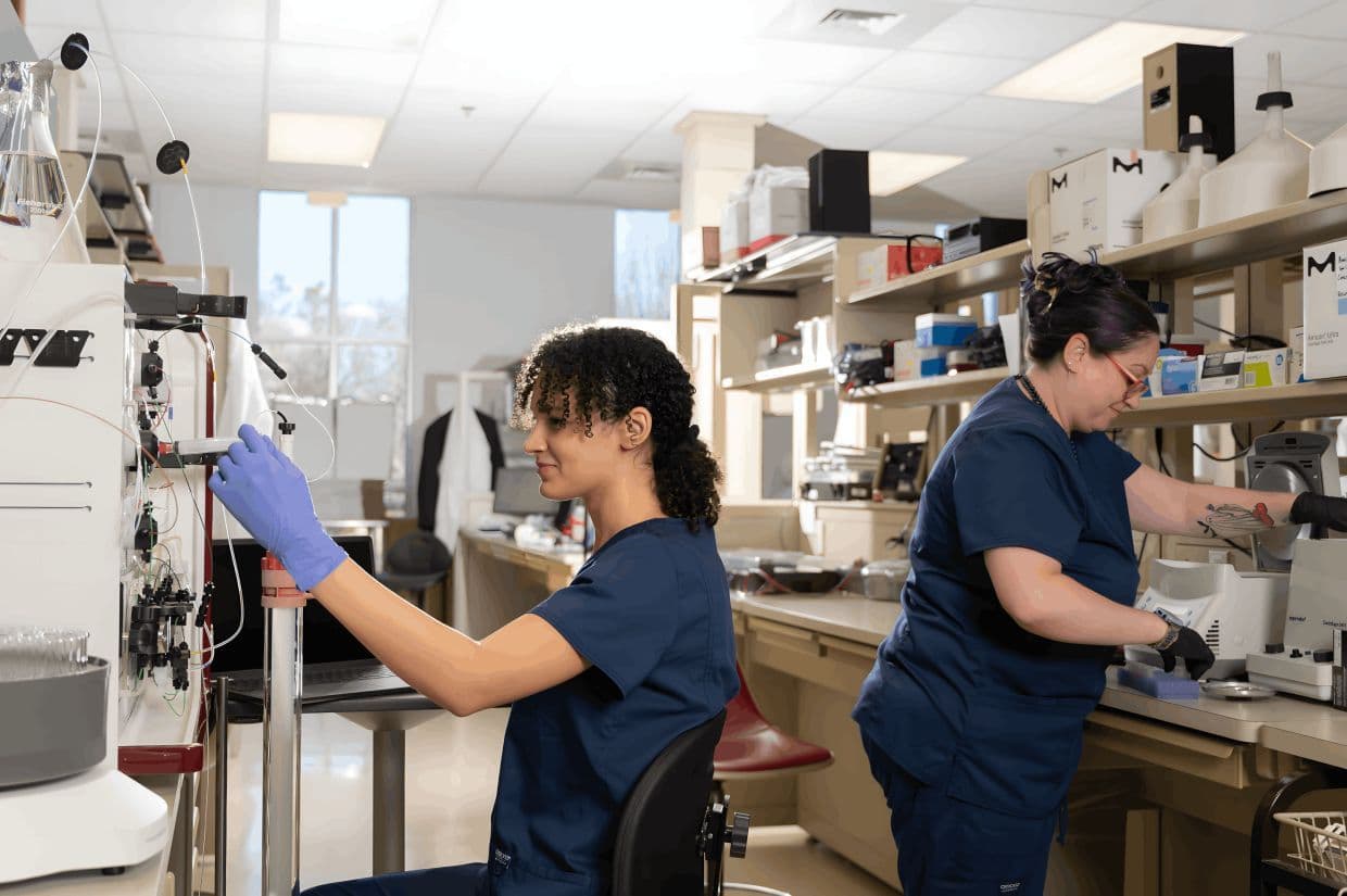 Two lab technicians from Athens Bioscience in navy scrubs and gloves work with equipment at their stations in a bright laboratory.