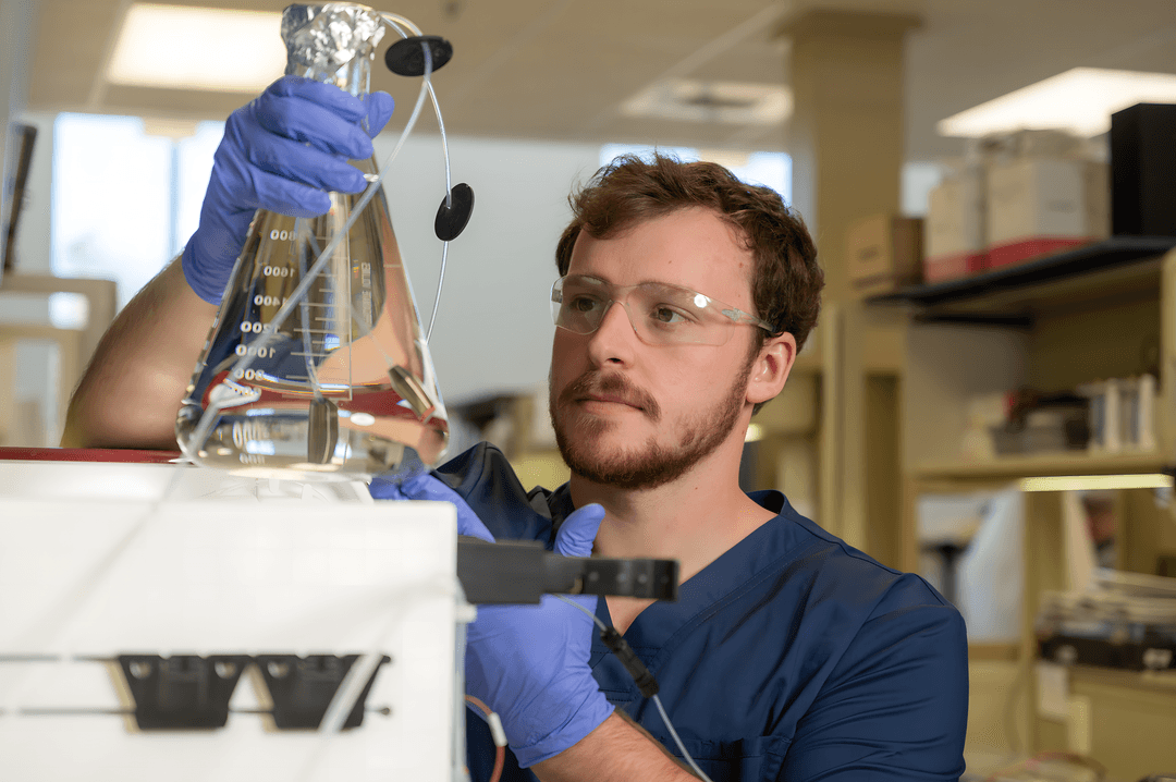 Lab technician from Athens Bioscience in safety glasses and blue gloves examines a glass flask in a laboratory setting.