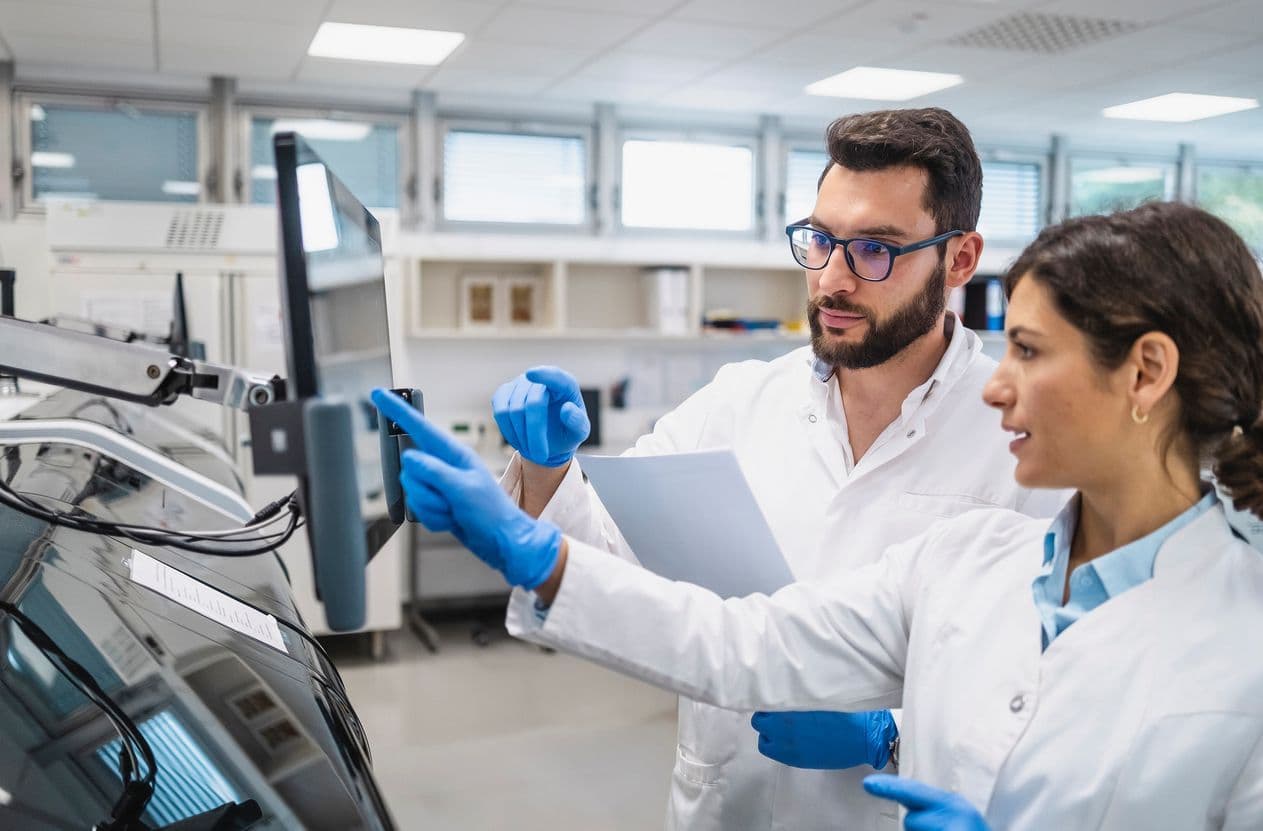 Two scientists in lab coats and blue gloves review data on an instrument display while holding documentation.