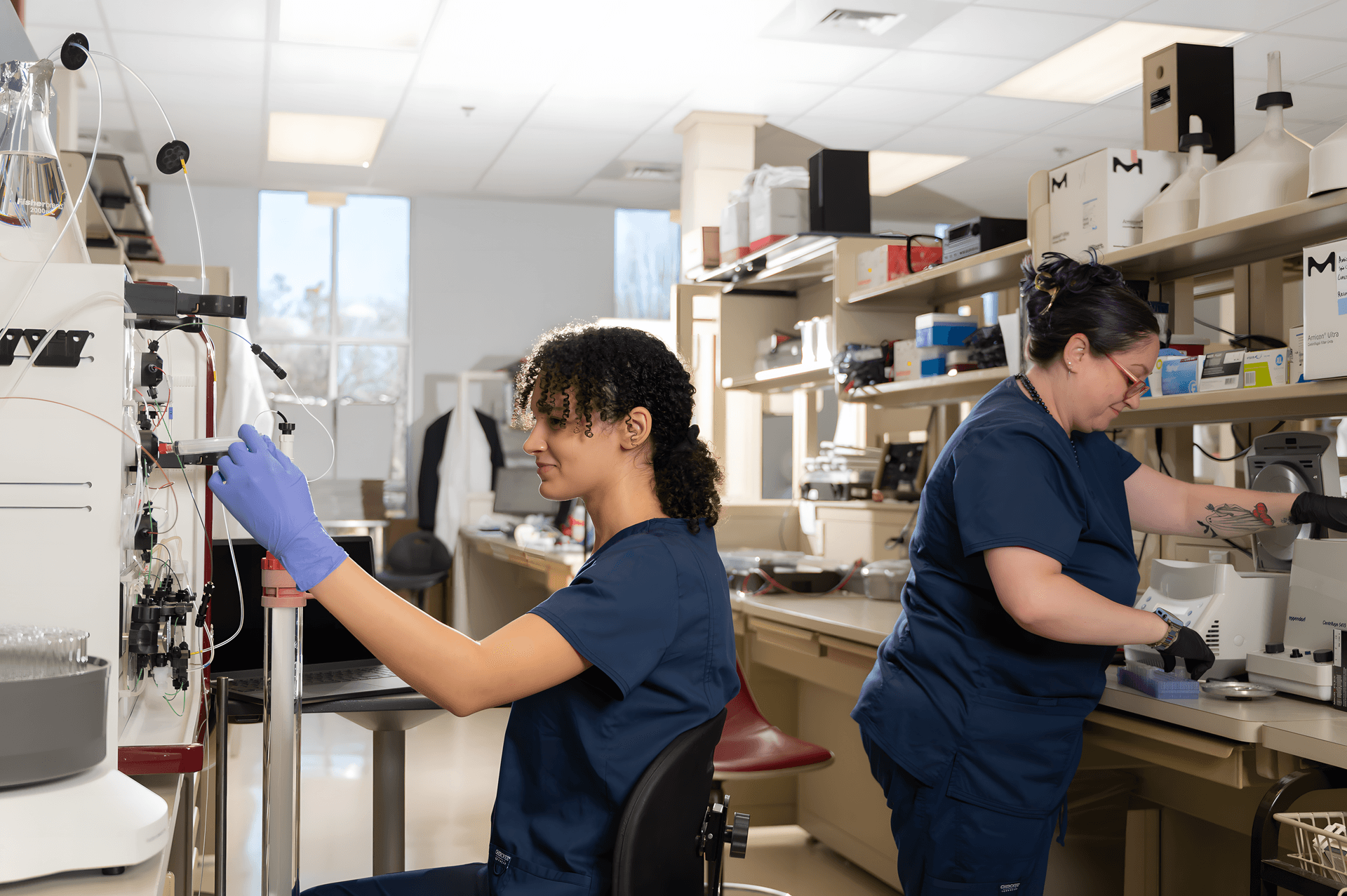 Two lab technicians from Athens Bioscience in navy scrubs and gloves work with equipment at their stations in a bright laboratory.